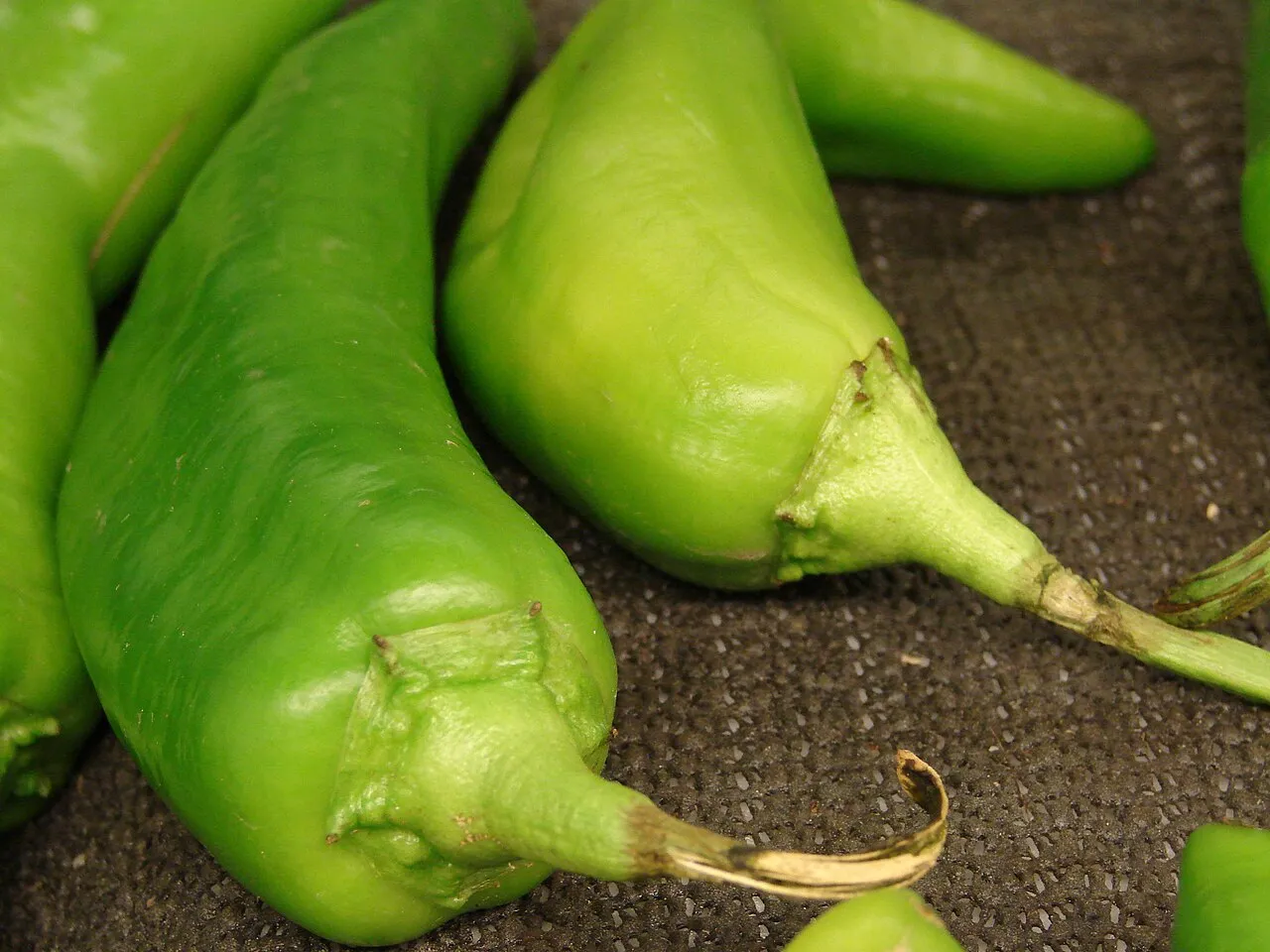 Several long tapering bright green Anaheim chile peppers with slightly wrinkled shoulders and thick green stems, photographed close up.