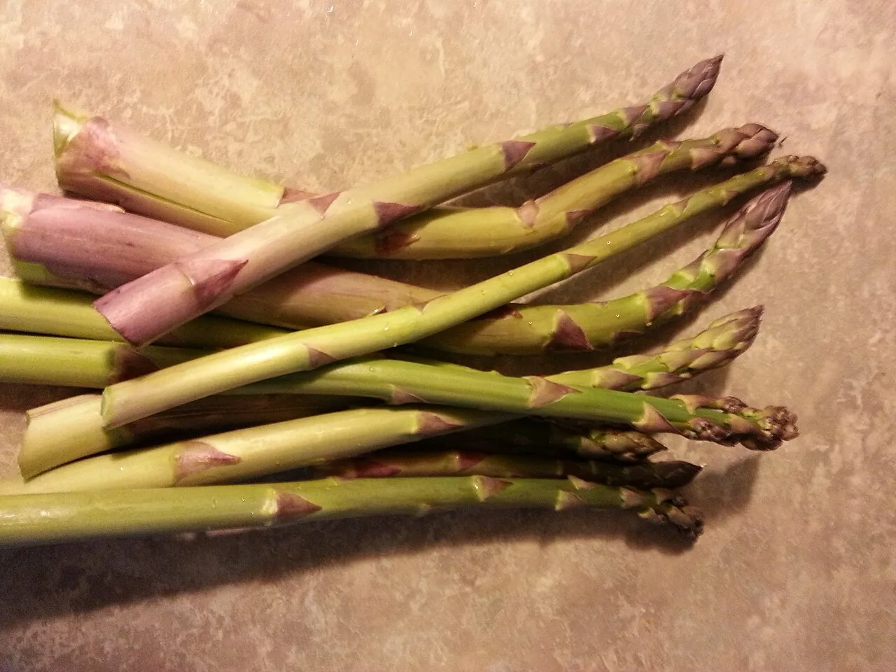 Freshly cut green asparagus spears laid on a counter, showing tight scaled tips and purple-tinged bracts along the stems.