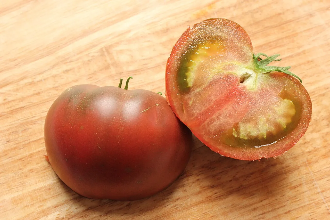 A whole Black Krim tomato with dark maroon-brown shoulders shading to red, beside a cut half showing dark red flesh and green gel around the seeds.